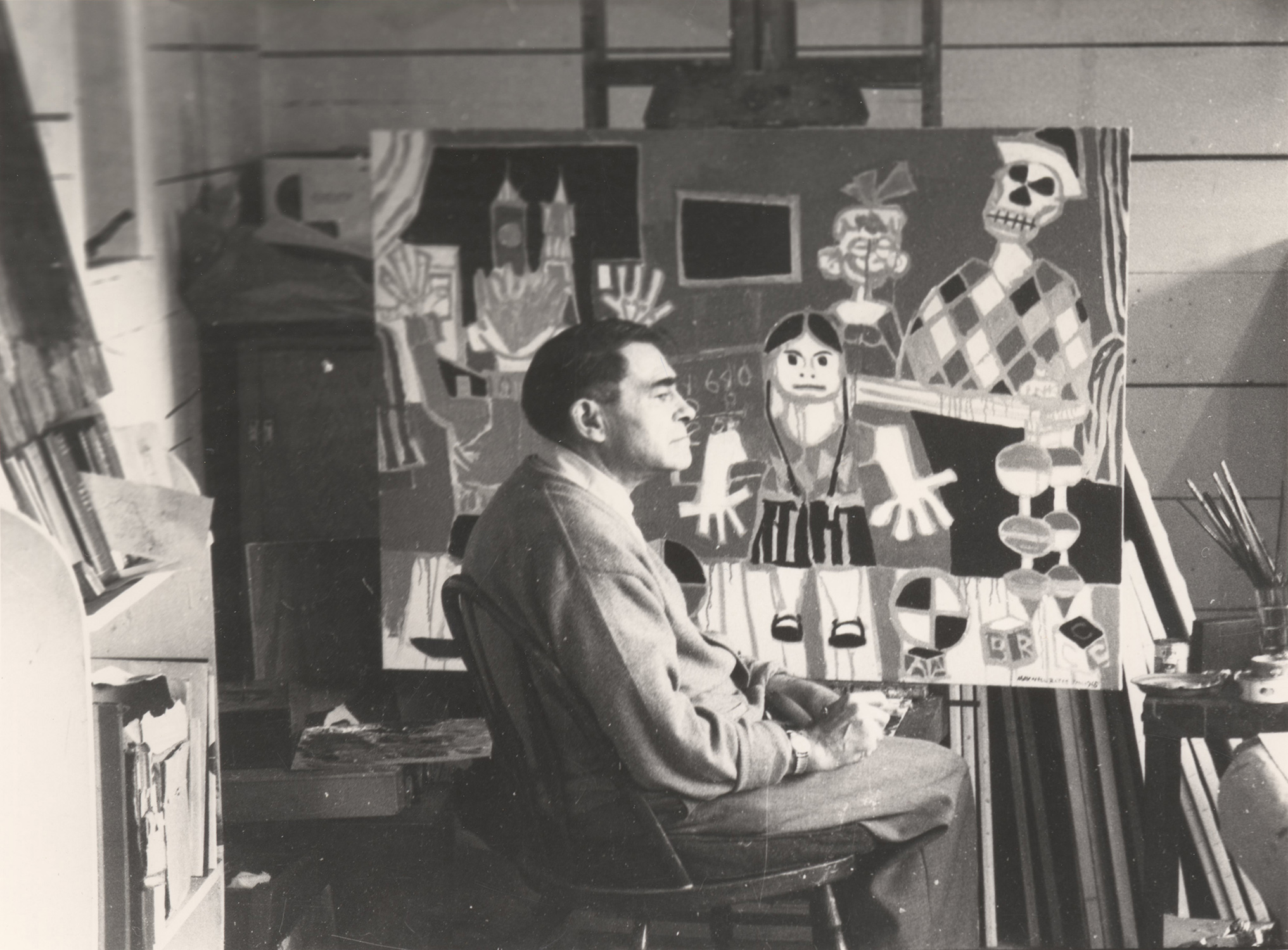Black and white photo of artist Maxwell Bates in his studio, he sits in front of his painting Kindergarten, facing right